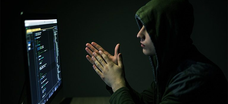 Black Hat Hacker staring at computer screen wearing hoodie as the room is illuminated by green light. White Hat Hacker conceptual.